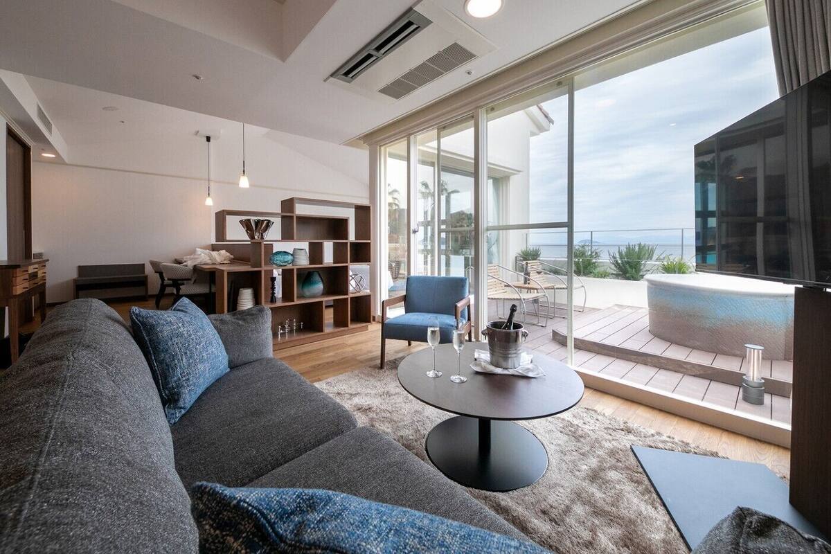 A modern living room with a large sofa, a coffee table, and a bookshelf. There is a balcony with a view of the ocean.