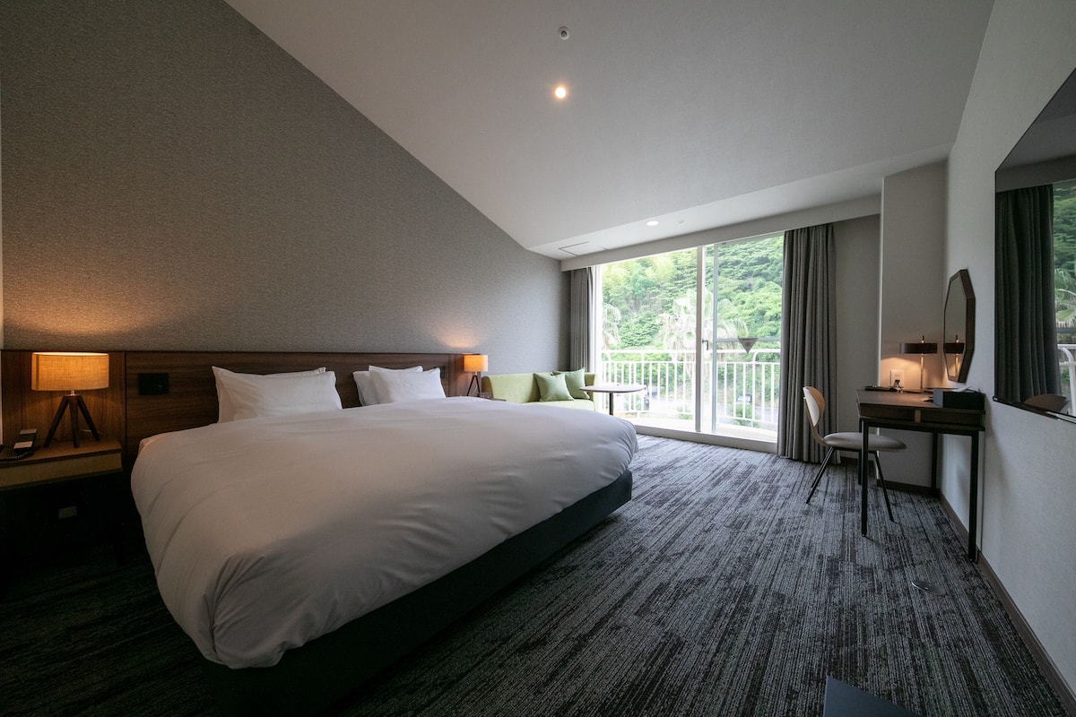 A hotel room with a large bed, a desk, a chair, and a balcony with a view of greenery.