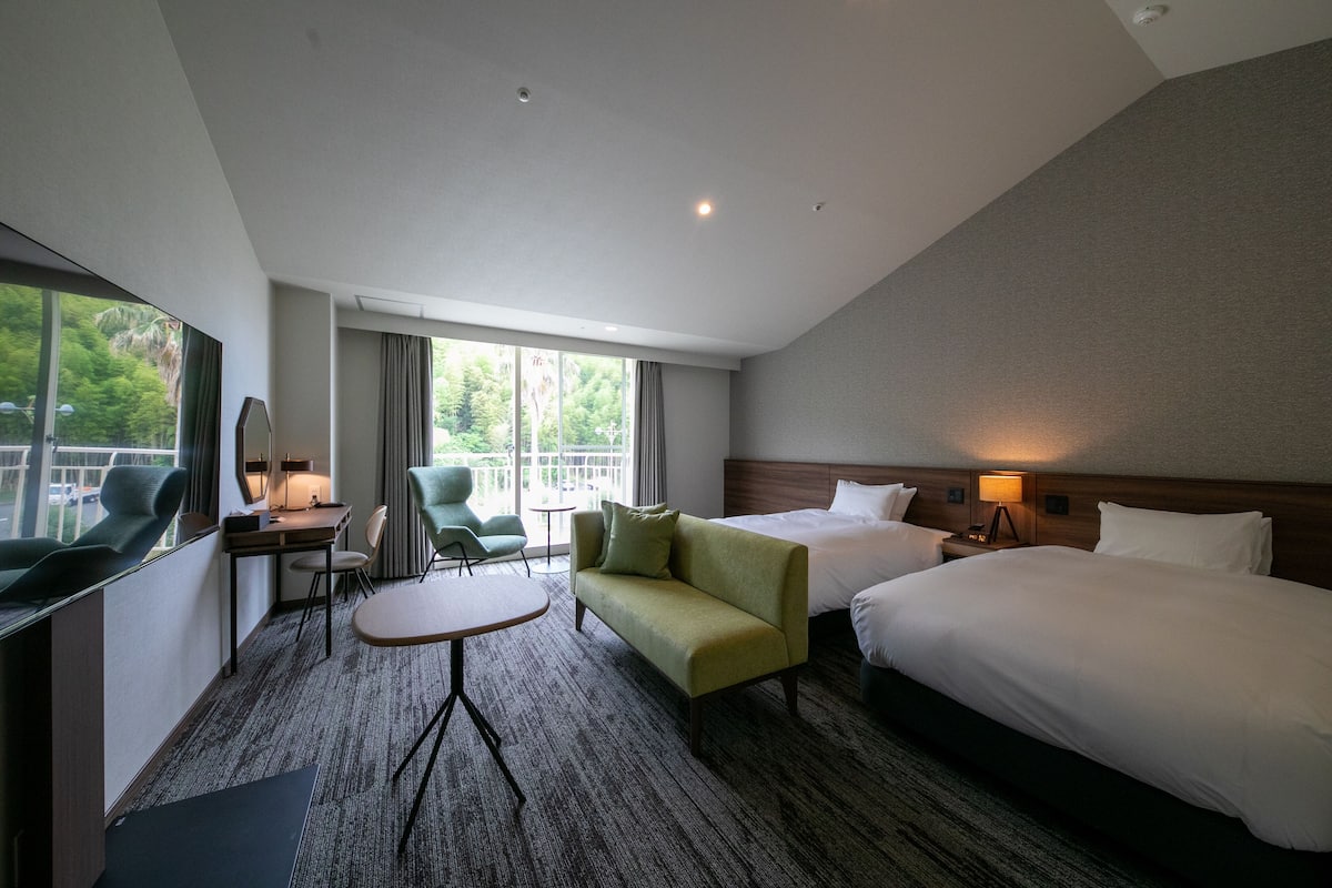 A hotel room with a large bed, a sofa, a desk, and a chair. There is a window with a view of trees.