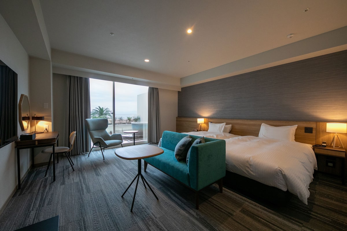 A hotel room with a bed, a sofa, a small table, a chair, and a desk. There is a window with curtains and a view of the outdoors.
