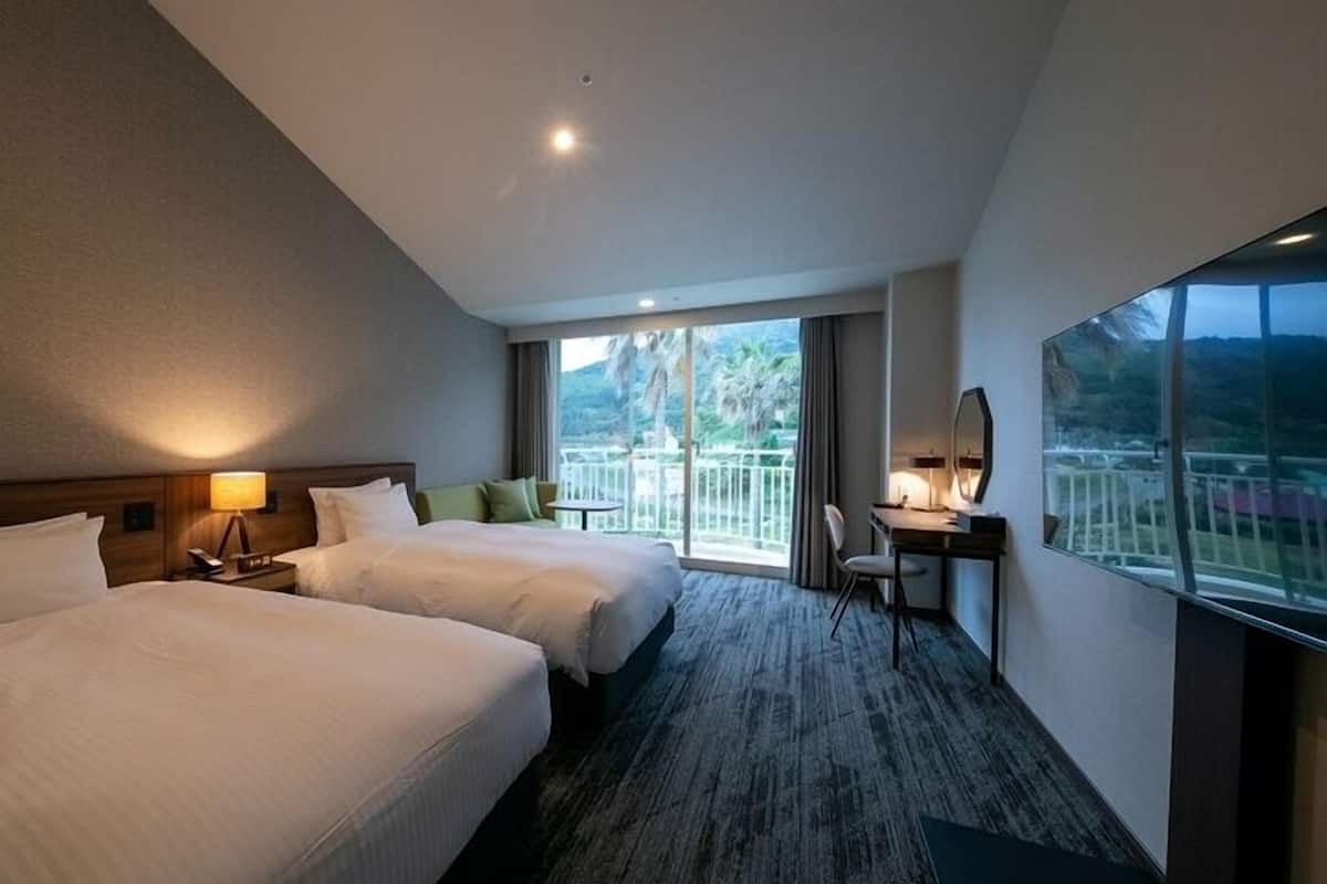 A hotel room with two beds, a desk, a chair, a balcony with a view, and a large window.