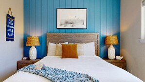 Ein Schlafzimmer mit hölzernem Kopfteil, zwei Nachttischlampen, einem gerahmten Bild über dem Bett und einer blauen Wand mit dem Schriftzug 'LET YOUR DREAMS SET Sail'.