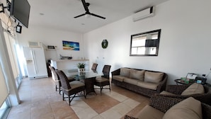 Uma sala de estar moderna com uma mesa de jantar de vidro, móveis de vime, um ventilador de teto e uma cozinha ao fundo.