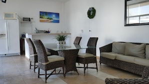 Uma área de refeições com cadeiras de vime e uma mesa de vidro, um sofá, uma cozinha com micro-ondas e um quadro com uma cena de praia.