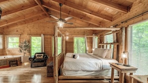 Une chambre dans un chalet en rondins, avec un lit à baldaquin, un ventilateur de plafond en bois, une table de chevet avec une lampe, et une vue sur l’extérieur grâce à de grandes fenêtres.