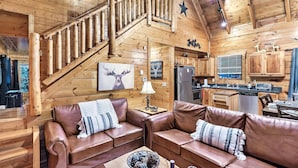 L’intérieur chaleureux d’un chalet avec des murs en bois, un escalier, un espace de vie doté de canapés en cuir, une table à manger et un coin cuisine.