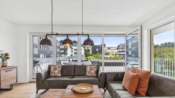 Een moderne woonkamer met een grijze bank, een houten salontafel en een uitzicht op woongebouwen door grote ramen.