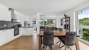 Een moderne keuken met witte kasten, een houten eettafel en zwarte stoelen. Er is uitzicht op een balkon met groen en gebouwen op de achtergrond.