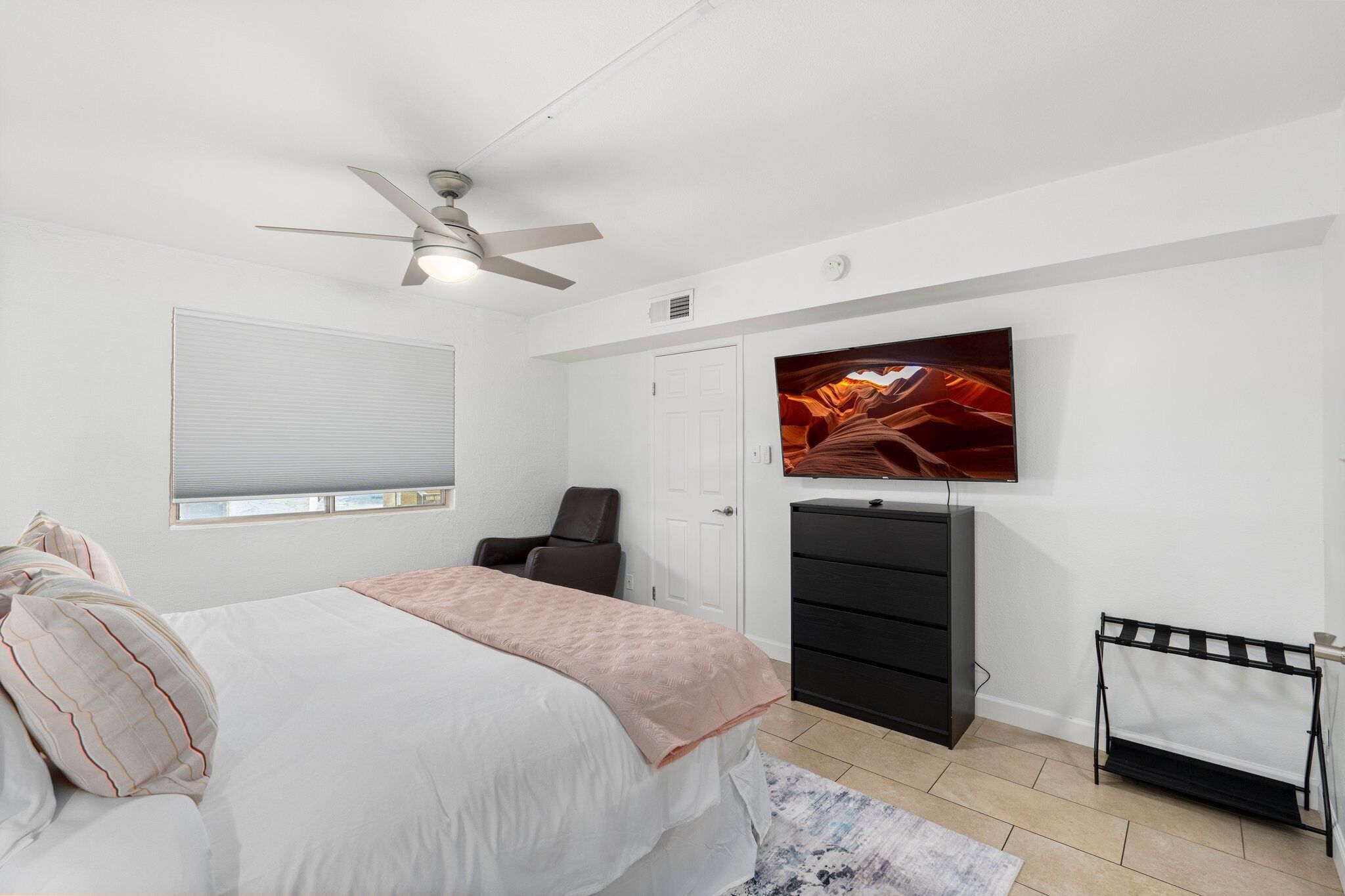 A bedroom with a bed, a ceiling fan, a TV on the wall, a chair, a dresser, and a radiator.