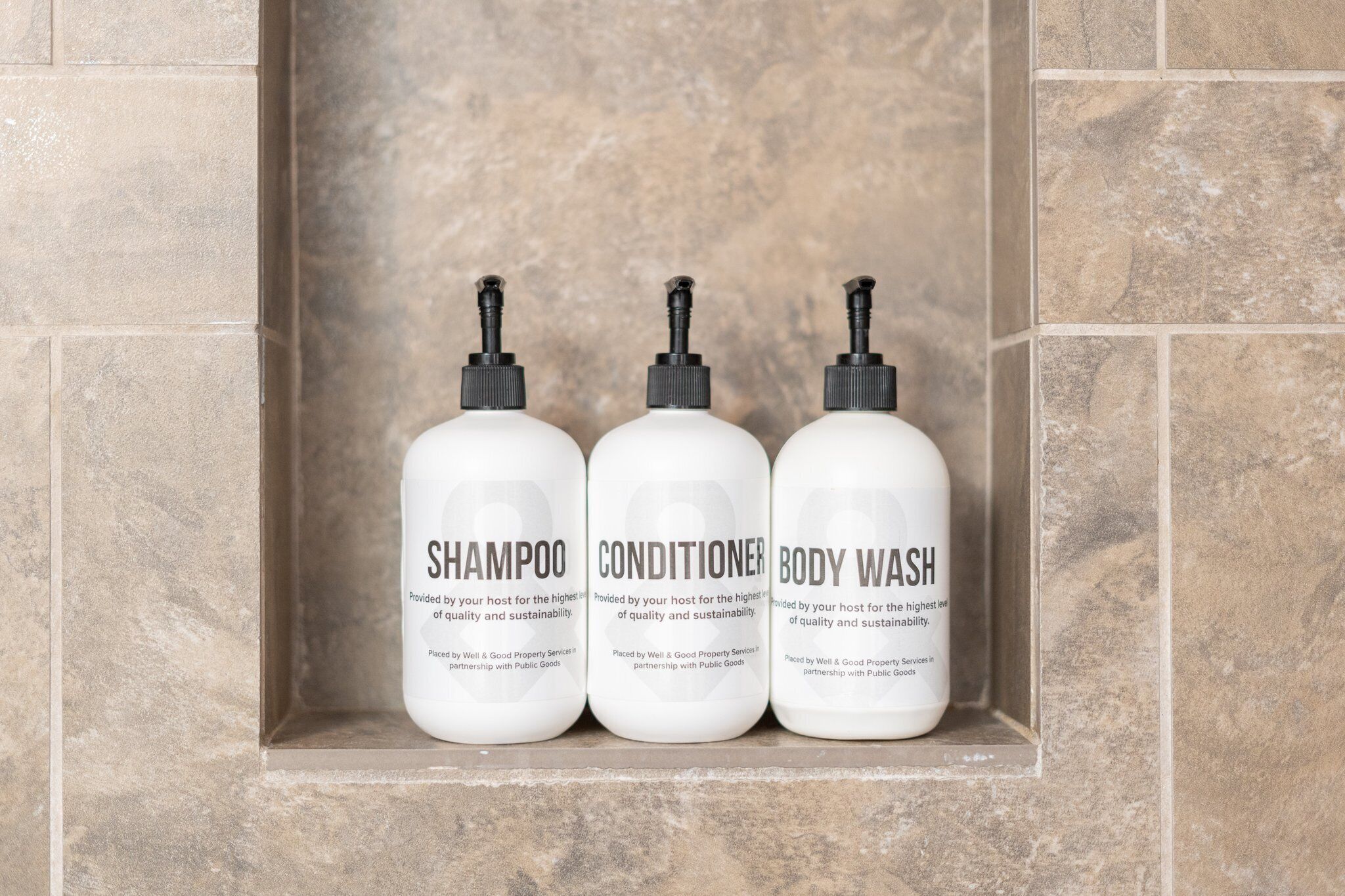 Three white bottles of shampoo, conditioner, and body wash in a tiled shelf.