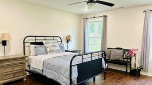 Une chambre à coucher comprenant un lit, des tables de chevet, un ventilateur de plafond, une fenêtre avec des rideaux et un banc agrémenté d’une plante.