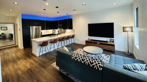 Una cocina moderna con una isla central, taburetes de bar, un televisor de pantalla plana grande y una zona de estar con sofá y reposapiés.