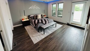 Un dormitorio moderno con cama, mesitas de noche, ventilador de techo y ventanas amplias.