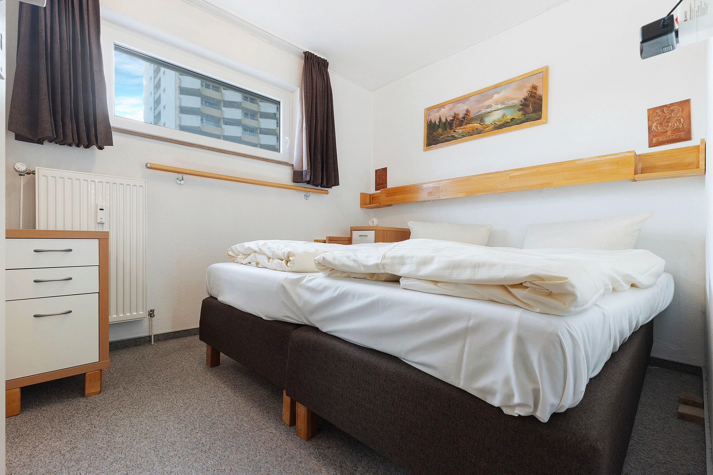 Een slaapkamer met een bed, een kaptafel, een raam met gordijnen, een radiator en een schilderij aan de muur.