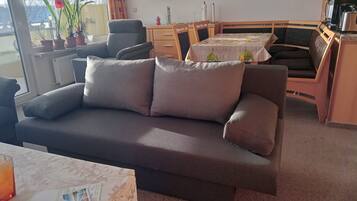 Een woonkamer met een bank, een eettafel, stoelen, een radiator, een raam met gordijnen, en een potplant.