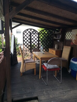 A covered outdoor seating area with a wooden table and chairs, a wicker bench, and a trash bin.