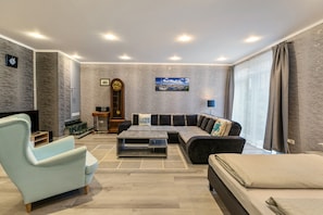 A modern living room with a sectional sofa, a light armchair, a coffee table, a TV, a clock, and a large window with curtains.