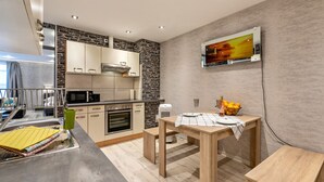 Une cuisine moderne avec un pare-feu en pierre, un téléviseur à écran plat fixé au mur, une table à manger avec un set de table et un panier de fruits posé sur la table.