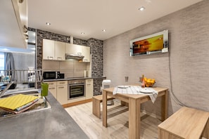 A modern kitchen with a stone backsplash, a flat-screen TV mounted on the wall, a dining table with a placemat, and a fruit bowl on the table.