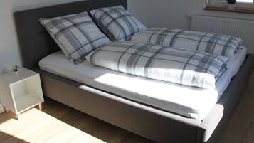 A bed with a grey headboard, white bedding, and two pillows. There is a small bedside table with a plant on it.