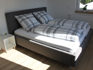 Una cama con cabecera gris, ropa de cama blanca y dos almohadas. Hay una mesita de noche pequeña con una planta encima.