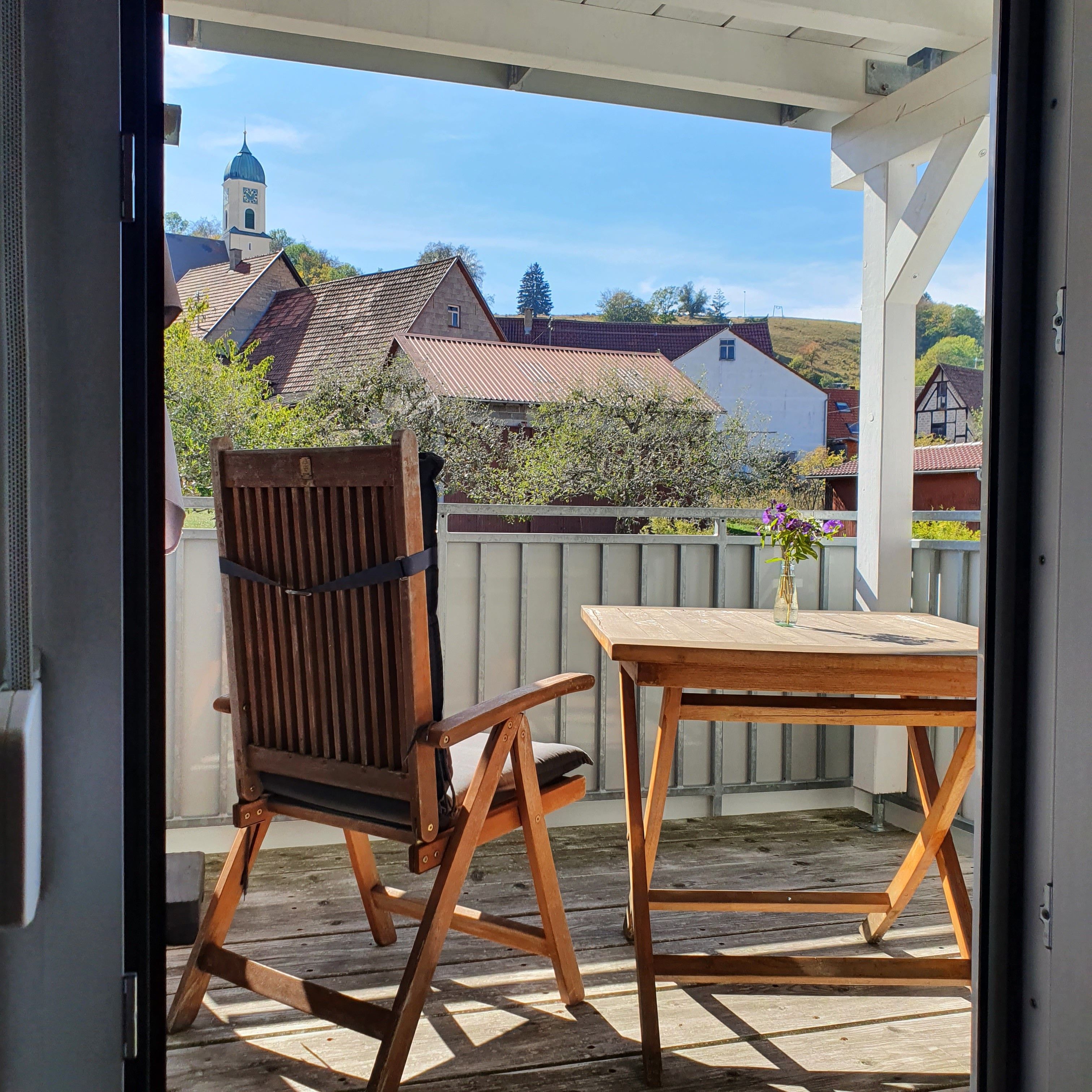 Ein Holztisch und ein Holzstuhl auf einer Holzterrasse mit Blick auf Häuser und ein Kirchturm.