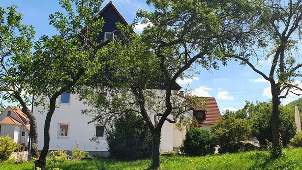 Une maison avec un toit en pente, entourée d’une végétation luxuriante et d’arbres.