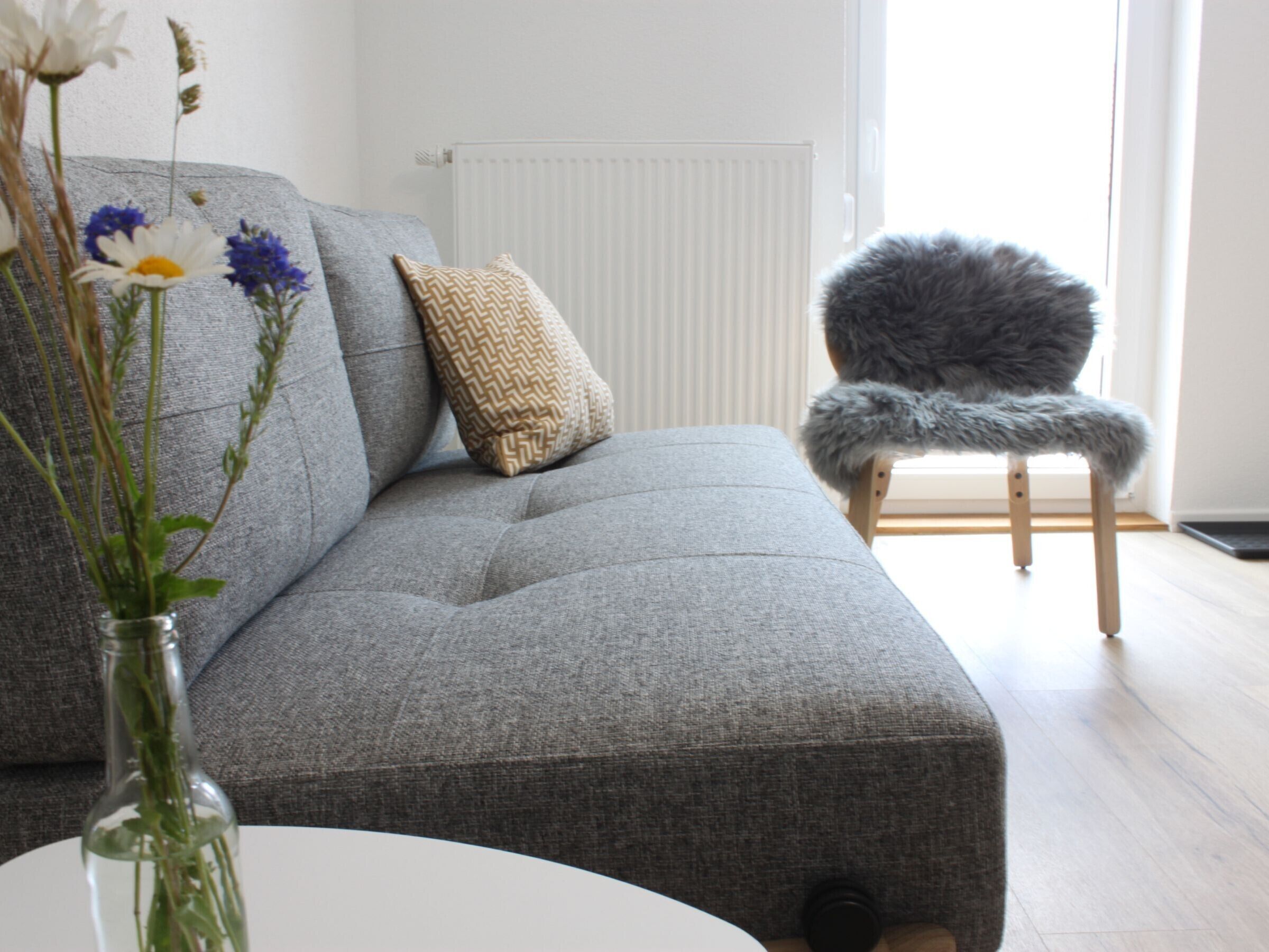A grey sofa with a patterned pillow, a fluffy chair, and a vase with flowers.
