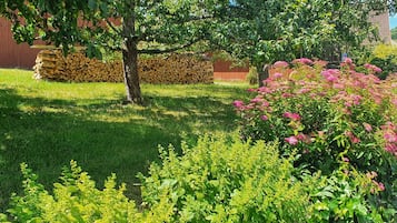 Een tuin met een boom, een stapel houtblokken en bloeiende planten.