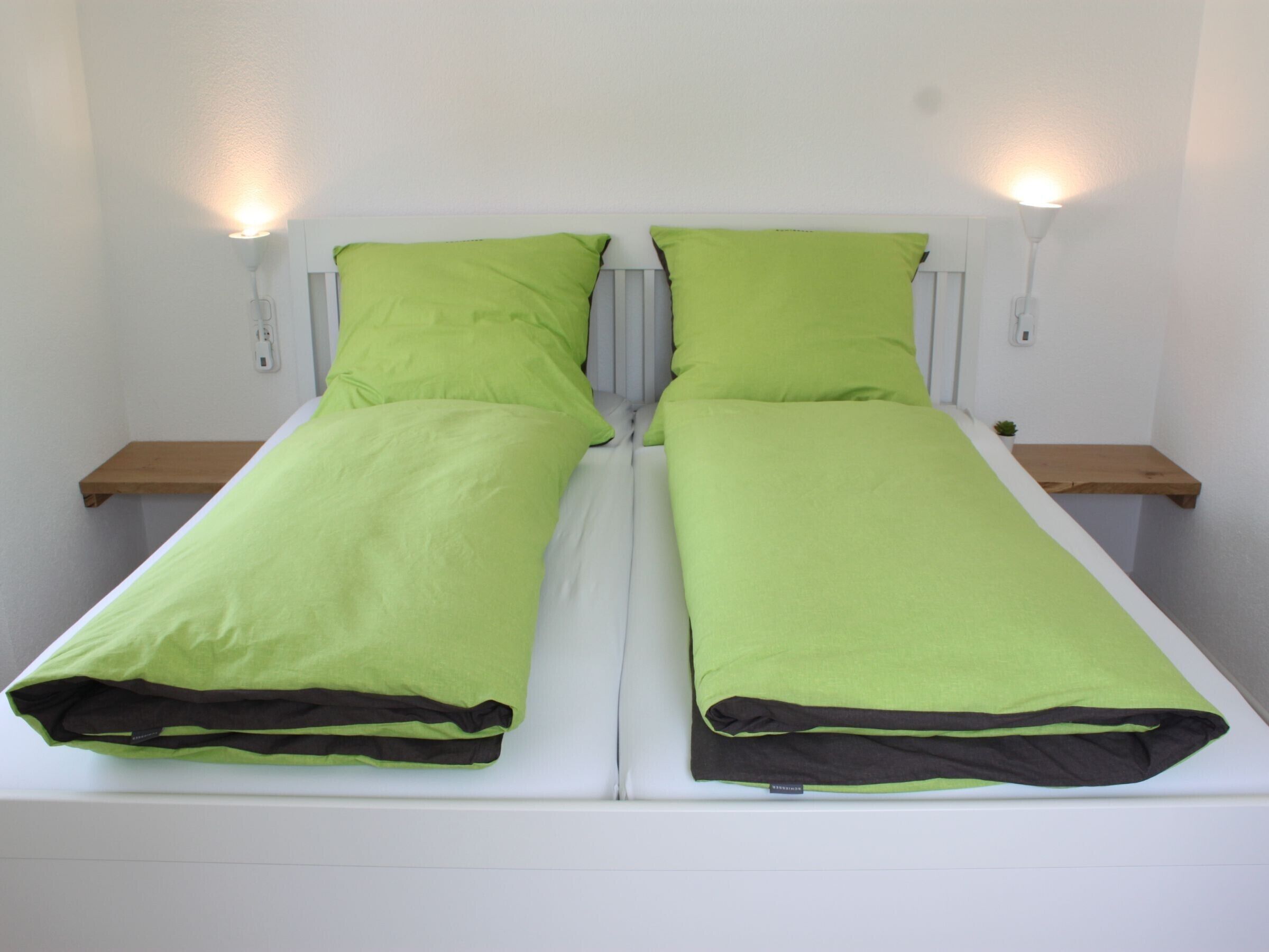 Un lit simple avec une literie verte, deux tables de chevet et des lampes de lecture fixées au mur.