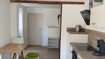 A compact kitchen with a wooden table, two green cushioned stools, a coffee machine, and a kettle.