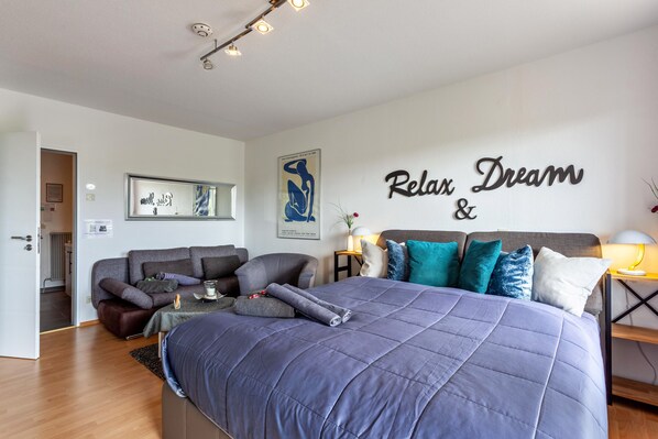 A bedroom with a large bed, a sofa, a bedside table with a lamp, and a wall with 'Relax Dream &' text.