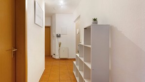 Un pasillo con puerta de madera, paredes blancas, suelo de baldosas y un estante blanco con una planta encima.