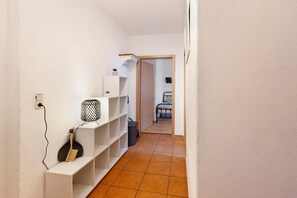 A narrow hallway with white walls and a tiled floor, leading to a room with a bed and a chair.