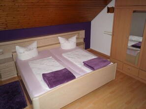 Makuuhuoneessa on puinen sängynpääty, valkoisilla lakanoiden peitetty sänky, violetti peitto ja peilillä varustettu puinen vaatekaappi.