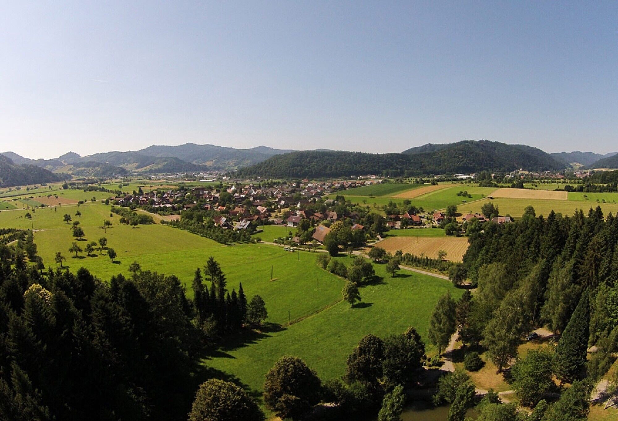 Un paisaje rural con un pequeño pueblo, campos verdes y zonas boscosas.