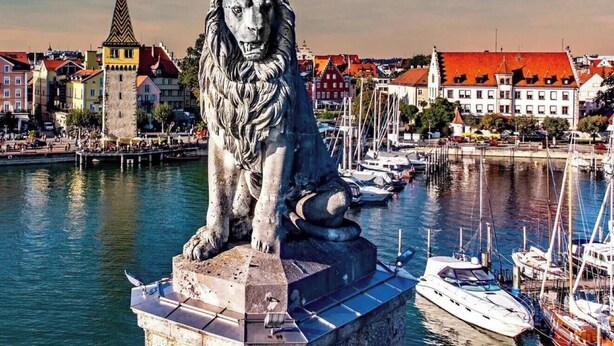 A statue of a lion on a pedestal by a waterfront with buildings and boats in the background.