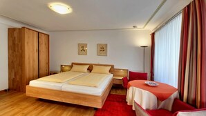 Quarto de hotel com uma cama de madeira, uma mesa pequena com toalha de mesa vermelha e uma cadeira vermelha.