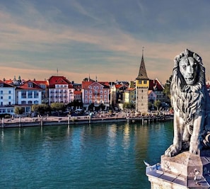 Une statue de lion, posée sur un piédestal, se trouve au bord de l’eau, avec des bâtiments et des bateaux en arrière-plan.