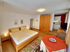 Une chambre avec un lit double, une petite table avec une assiette, une télévision et, au fond, un coin cuisine.