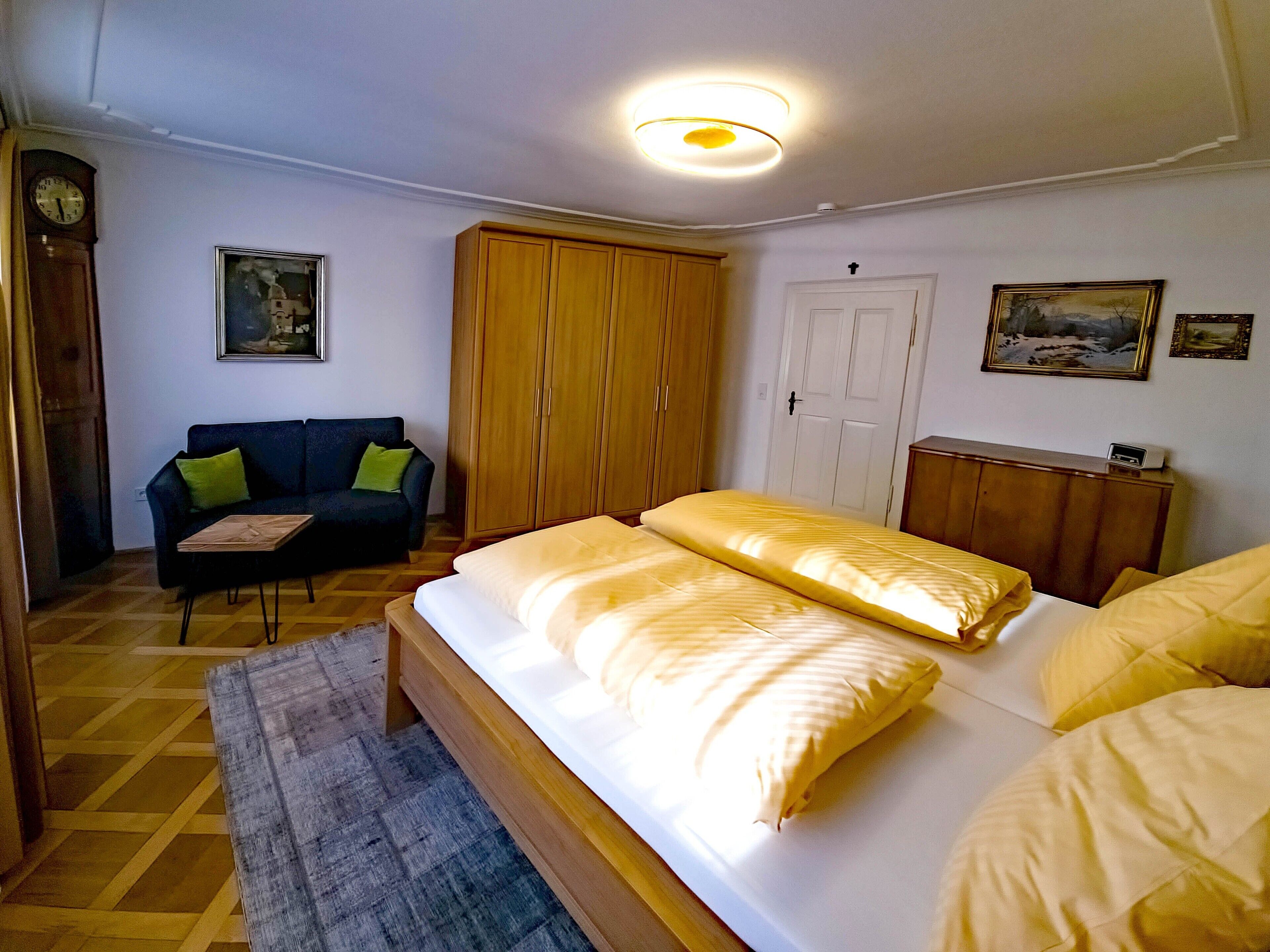 Ein Schlafzimmer mit einem Bett, einem Holzschrank, einem Nachttisch aus Holz, einem Holz-Kopfteil, einer Holztür, einem gerahmten Bild an der Wand und einer Wanduhr.