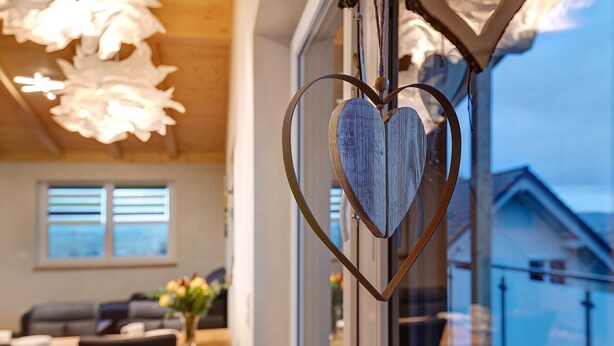 A heart-shaped metal ornament hanging near a window, a dining area with a table and chairs, and a living room with a sofa and a vase of flowers. - Panorama Suite Apartment with Balcony, Grill Hut and Infrared Cabin (Donaueschingen)