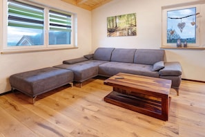 A modern living room with a sectional sofa, a wooden coffee table, and two windows with blinds.