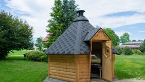 A wooden cabin with a sloped roof and a chimney, situated on a paved area with grass and trees around. - Panorama Suite Apartment with Balcony, Grill Hut and Infrared Cabin (Donaueschingen)