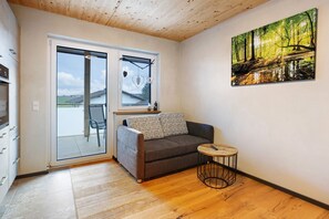 A room with a sliding glass door leading to a balcony, a grey sofa with patterned cushions, a small round table, and a painting of a forest scene on the wall. - Kuschel Apartment with Balcony, Barbecue Hut, and Infrared Cabin (Donaueschingen)