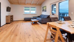 A spacious living area with a wooden ceiling, a sofa, a dining table with chairs, a TV, and a large window with a view.