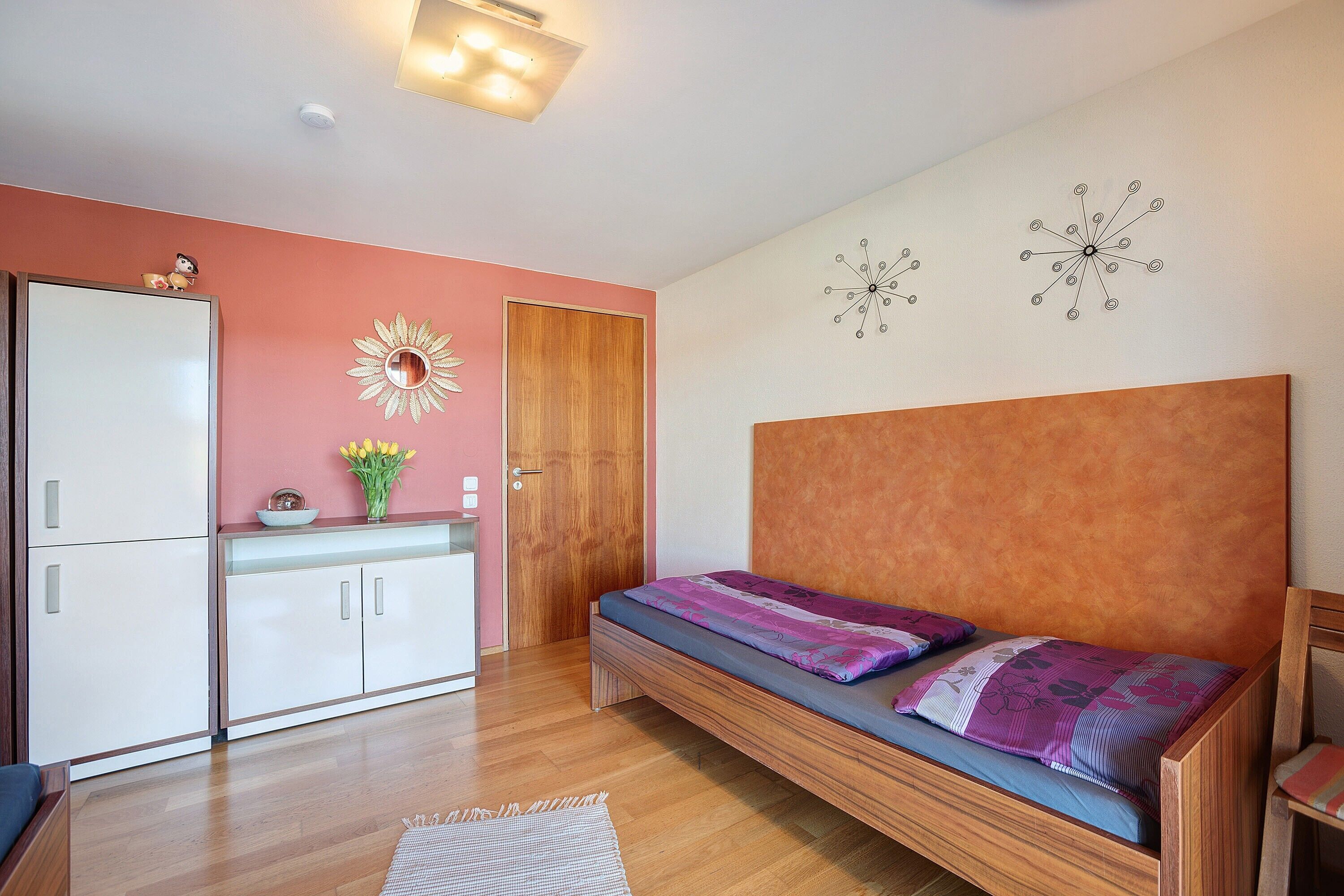 Een slaapkamer met een houten bed, een witte kledingkast, een roze muur en een houten deur.