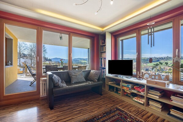 A modern living room with a sofa, TV, bookshelves, and large windows offering a view of the outdoors.