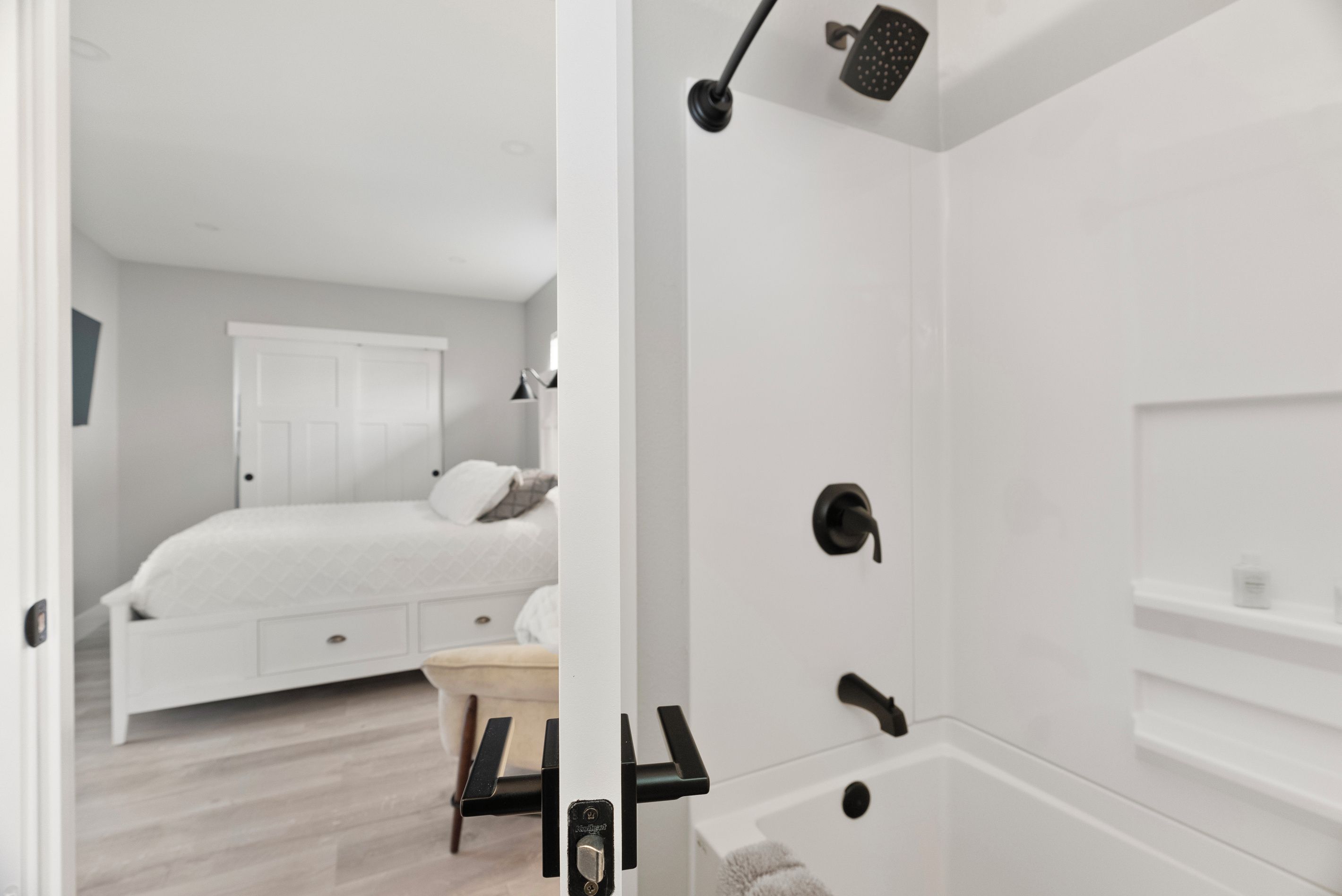 A modern bathroom with a bathtub, shower, and toilet. There is a bed in the adjacent room with a nightstand and a lamp.