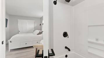 A modern bathroom with a bathtub, shower, and toilet. There is a bed in the adjacent room with a nightstand and a lamp.